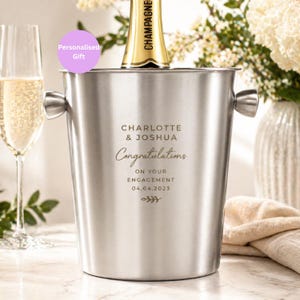 May include: A stainless steel champagne bucket with handles, holding a champagne bottle. The bucket is engraved with "CHARLOTTE & JOSHUA Congratulations ON YOUR ENGAGEMENT 04.04.2023". A glass of champagne is visible to the left.