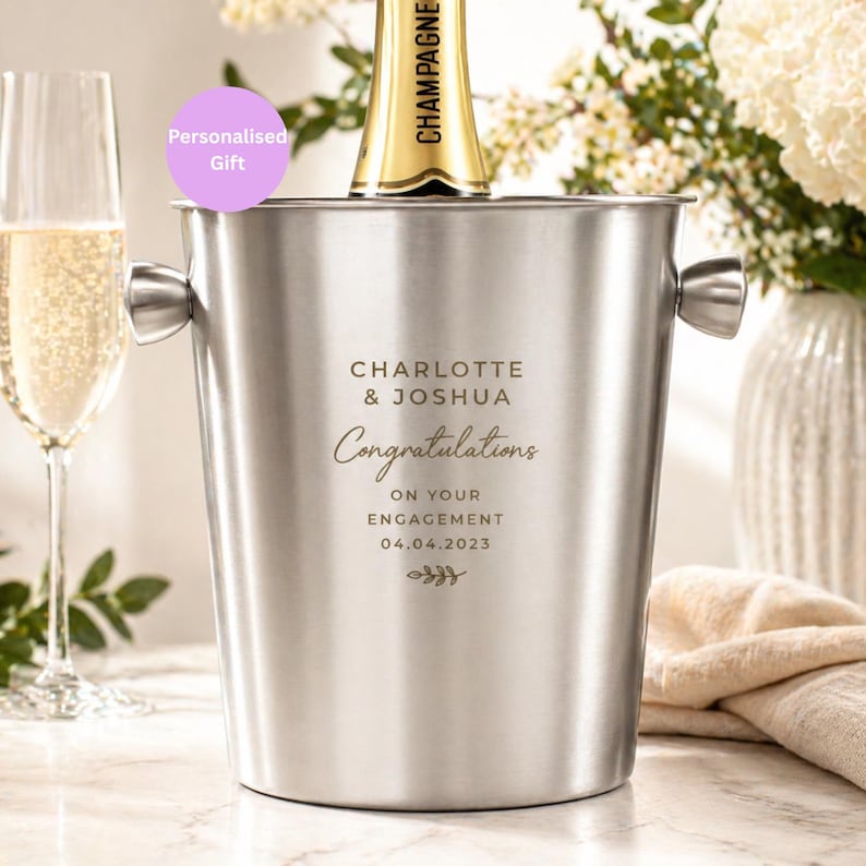 May include: A stainless steel champagne bucket with handles, holding a champagne bottle. The bucket is engraved with "CHARLOTTE & JOSHUA Congratulations ON YOUR ENGAGEMENT 04.04.2023". A glass of champagne is visible to the left.