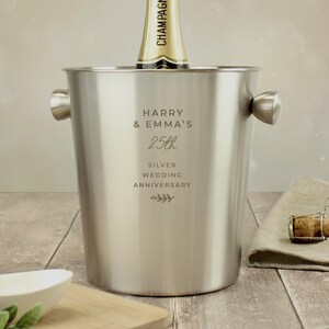 May include: A stainless steel champagne bucket with two handles, holding a champagne bottle. The bucket is engraved with "HARRY & EMMA'S 25th SILVER WEDDING ANNIVERSARY". A cork and napkin are on the wooden surface.