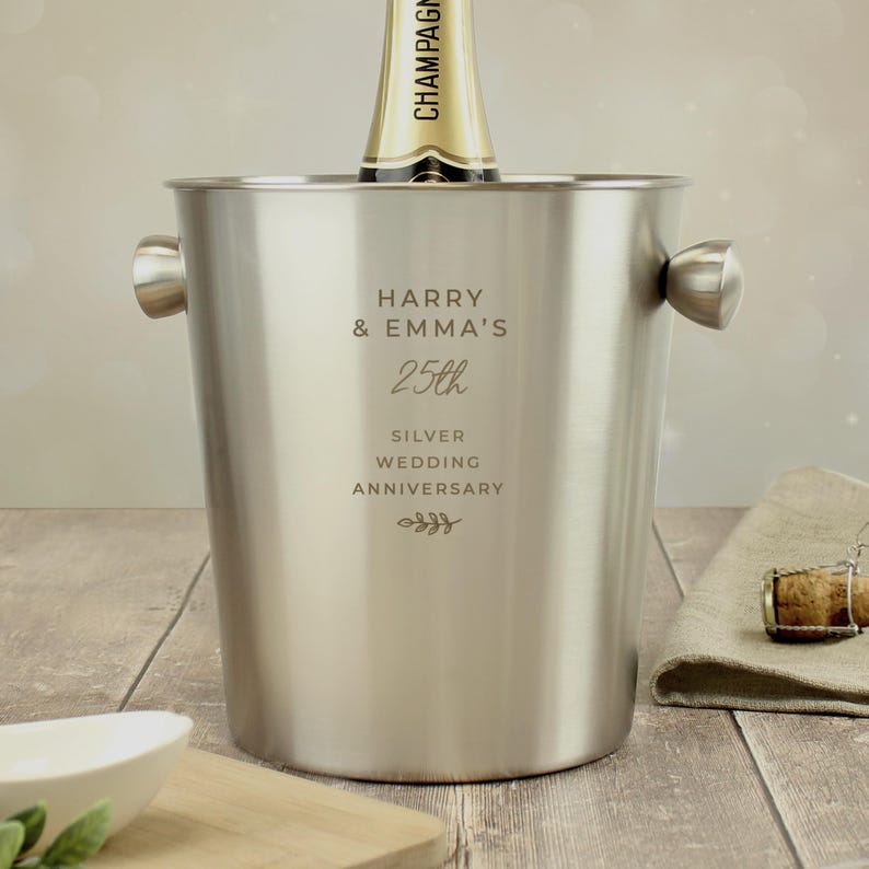 May include: A stainless steel champagne bucket with two handles, holding a champagne bottle. The bucket is engraved with "HARRY & EMMA'S 25th SILVER WEDDING ANNIVERSARY". A cork and napkin are on the wooden surface.