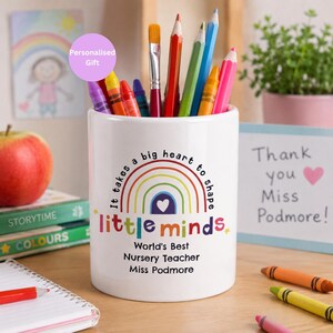 May include: White ceramic pencil pot with a rainbow design and the text "It takes a big heart to shape little minds." The pot is filled with colourful pencils, crayons, and a paintbrush. Text on the pot reads "World's Best Nursery Teacher Miss Podmore."
