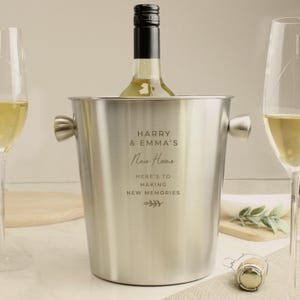 May include: A stainless steel wine bucket with handles, holding a bottle of white wine. The bucket is engraved with the text "HARRY & EMMA'S New Home" and "HERE'S TO MAKING NEW MEMORIES". Two champagne flutes are on either side.