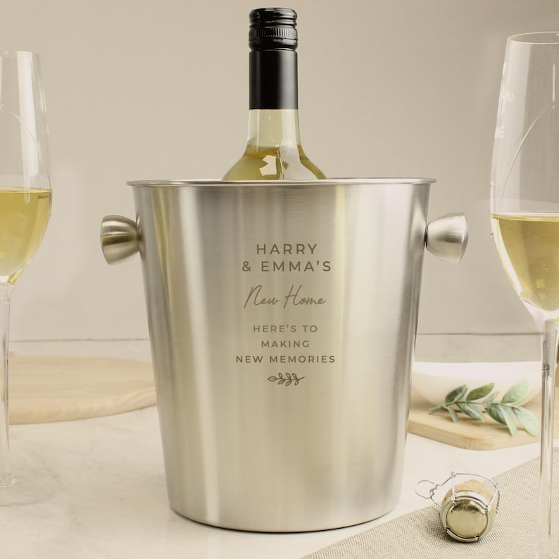 May include: A stainless steel wine bucket with handles, holding a bottle of white wine. The bucket is engraved with the text "HARRY & EMMA'S New Home" and "HERE'S TO MAKING NEW MEMORIES". Two champagne flutes are on either side.