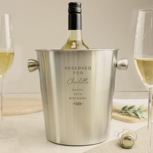 May include: A stainless steel wine bucket with handles, holding a bottle of white wine. The bucket is engraved with "RESERVED FOR Charlotte HAPPY 30TH BIRTHDAY" and a decorative sprig. A champagne cork rests nearby.