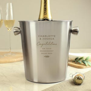 May include: A stainless steel champagne bucket with handles, holding a champagne bottle. The bucket is engraved with the names "Charlotte & Joshua" and the word "Congratulations" along with the date "04.04.2023".