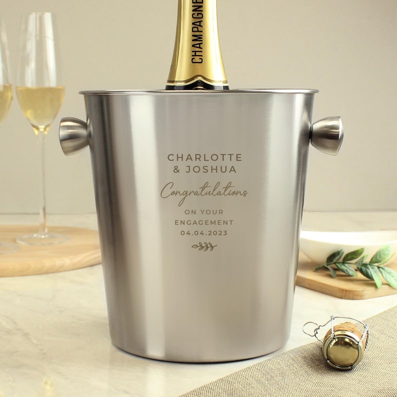 May include: A stainless steel champagne bucket with handles, holding a champagne bottle. The bucket is engraved with the names "Charlotte & Joshua" and the word "Congratulations" along with the date "04.04.2023".