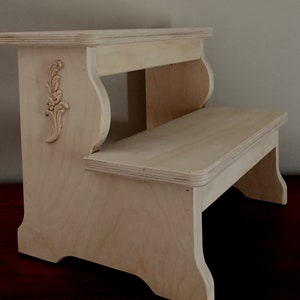 May include: A natural wood two-step stool with a decorative floral carving on the side. The stool has a rounded top step and a rectangular bottom step.