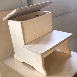 May include: A wooden step stool with a hinged lid that opens to reveal a storage compartment. The stool is made of light-colored wood and has a simple, modern design.