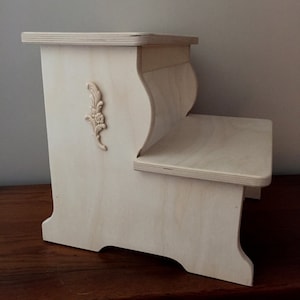 May include: A white wooden two-step stool with a decorative floral design on the side. The stool has a rounded top step and a rectangular bottom step.
