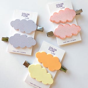 Op de afbeelding: Vier haarclips in de vorm van wolken in pastelkleuren: lavendel, roze, geel en oranje. Elke clip zit op een witte kaart met de tekst "COLOR FLOW LAB" en "made with in Berlin". De clips hebben zilveren metalen sluitingen.