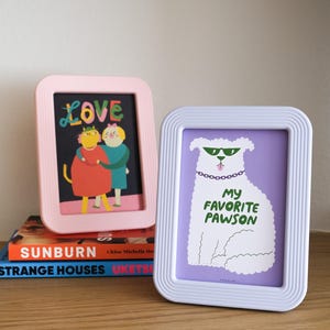 May include: Two rectangular picture frames with ribbed borders, one light pink and one white. The pink frame holds an illustration of two figures with the word LOVE. The white frame holds a drawing of a white dog with green sunglasses and the text MY FAVORITE PAWSON.