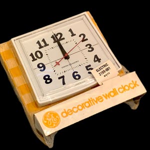 May include: A white square wall clock with black numerals and red second hand. The clock face has the words "GENERAL ELECTRIC" printed on it. The clock is packaged in a yellow and white checkered box with the words "decorative wall clock" printed on the packaging.