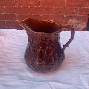 May include: A brown ceramic pitcher with a wide mouth and a handle. The pitcher is decorated with a raised design of people and trees.