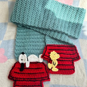 May include: A handmade crochet scarf in shades of teal, with two red and black Snoopy dog house appliques. One applique features Snoopy, the other features Woodstock. The scarf is made of soft yarn.