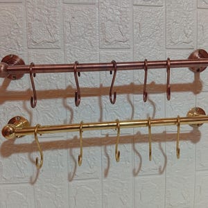 May include: Two metal kitchen utensil racks. The top rack is bronze, and the bottom rack is gold. Each rack has a horizontal bar with several curved hooks for hanging items. The racks are mounted on a textured, off-white wall.