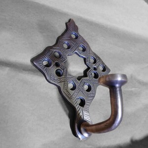 Könnte beinhalten: Ein dekorativer Haken aus Metall mit einem komplizierten geometrischen Muster. Der Haken ist aus Messing und hat eine dunkle Patina. Er ist eine einzigartige und stilvolle Ergänzung für jedes Zuhause.