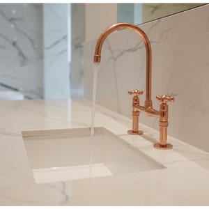 Pode incluir: Uma torneira de ponte em tom de cobre polido com dois manípulos em cruz montada numa bancada de mármore branco com veios cinzentos. A água flui do bico curvo para um lavatório retangular branco integrado na superfície de pedra.