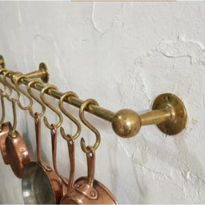 May include: A wall-mounted brass rail featuring a series of matching brass S-hooks. Several small copper pans with metallic handles are suspended from the hooks. The rail is secured to the wall with circular brass plates at each end, featuring a polished finish.
