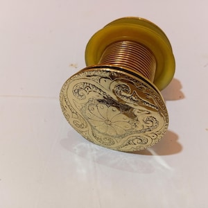 May include: A decorative bottle stopper with a gold-coloured, engraved design. The stopper features a floral pattern and a brass-coloured, threaded neck. The top is a translucent yellow colour.
