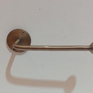 May include: A bronze-colored, wall-mounted toilet paper holder. The holder features a circular mounting plate, a curved arm, and a spherical end. The fixture is designed for easy access and dispensing of toilet paper rolls. The design is simple and functional.