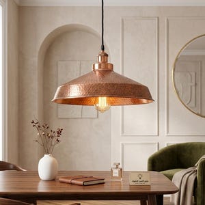 Puede incluir: Una lámpara colgante de color cobre con textura martillada cuelga sobre una mesa de madera. La lámpara tiene una pantalla cónica ancha y una bombilla visible de estilo Edison. La lámpara está suspendida de un cable negro, añadiendo un toque industrial al comedor.