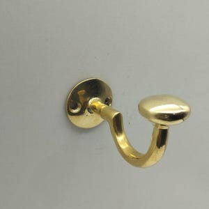 Könnte beinhalten: Ein goldfarbener Haken aus Metall mit abgerundeter Oberseite und einem gebogenen Haken. Der Haken ist an einer weißen Wand befestigt.