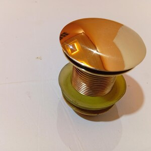 May include: A polished, gold-coloured sink drain stopper with a rounded top. The stopper has a threaded, copper-coloured body and a green rubber gasket. The drain stopper is designed to fit into a standard sink drain.