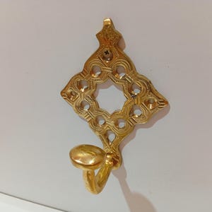 May include: A decorative brass wall hook with an ornate diamond-shaped design and a curved hook. The hook is gold-colored and features intricate cut-out patterns and a small hook for hanging items. The hook is designed to be mounted on a wall.