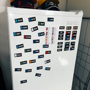 Puede incluir: Un refrigerador blanco con imanes que muestran los nombres de los pilotos de Fórmula 1, incluyendo RUS, ALB, LAW, GAS, BEA, ANT, HAM, TSU, DOO, PER, LEC, HUL, NOR, VER, ALO, STR y OCO. Los imanes son de varios colores, incluyendo azul, rojo, verde y negro.