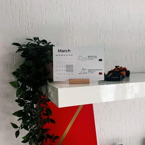 Könnte beinhalten: Ein weißer Kalender mit dem Monat März. Der Kalender zeigt den Albert Park Circuit in Melbourne, Australien und den Shanghai International Circuit in Shanghai, China. Der Kalender steht auf einem weißen Regal mit einem roten Buch und einer kleinen Topfpflanze.