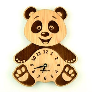 Puede incluir: Un reloj de madera con forma de panda con una esfera en blanco y negro. El reloj tiene una cara de panda en blanco y negro con una boca sonriente y ojos negros. La esfera del reloj tiene números y manecillas negros. El reloj está hecho de madera y tiene un acabado de madera natural.