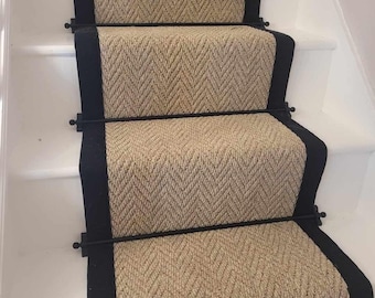 May include: A staircase runner featuring a tan herringbone pattern with thick black borders. The runner is secured to white wooden stairs with horizontal black metal stair rods that span the width of the carpeted section on each step.