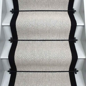 May include: A staircase runner featuring a light gray and white herringbone pattern with solid black borders on each side. The runner is secured to the white wooden stairs with thin, horizontal black metal rods that span the width of the carpet.