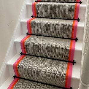 May include: Staircase featuring chevron patterned carpet runners in grey and white. The runners have a pink border with an orange stripe. Black metal rods secure the runners to the white stairs.