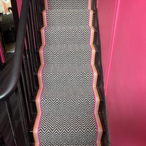 May include: Black and white herringbone patterned carpet runner with a pink, orange, and gray border. The runner is on a set of stairs with a black railing.