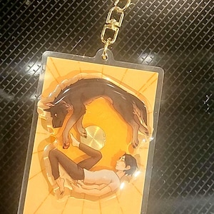 May include: A clear acrylic keyring featuring an illustration of a black dog and a person on a yellow background with sun rays. The keyring has a gold-coloured chain and clasp.