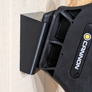 May include: A black, angular bracket is mounted on a light-colored wooden surface. The bracket is connected to a black, textured handle with the brand name "CANNON" printed in white.