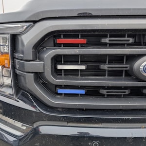 Può includere: Una griglia anteriore di un veicolo nera con tre inserti rettangolari orizzontali rossi, bianchi e blu, posizionati all'interno delle lamelle in plastica grigio scuro accanto a un emblema Ford e un faro trasparente con luce laterale arancione.