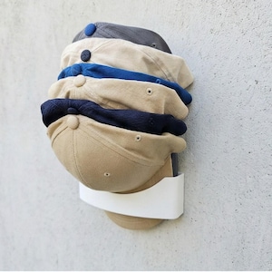 Könnte beinhalten: Eine Reihe von Baseballkappen in verschiedenen Farben, wie Beige, Marineblau und Blau, sind auf einem weißen, an der Wand befestigten Halter gestapelt. Die Kappen sind übereinander angeordnet und zeigen ihre unterschiedlichen Farben und Stile.