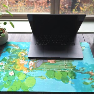 Könnte beinhalten: Ein Laptop liegt auf einem lebendigen Mauspads, das eine Frau mit langen blonden Haaren zeigt, die auf einem Bambusfloß inmitten von Seerosen und Koi-Karpfen treibt. Das Mauspad ist überwiegend blau und grün, mit einer detaillierten Wasserszene.