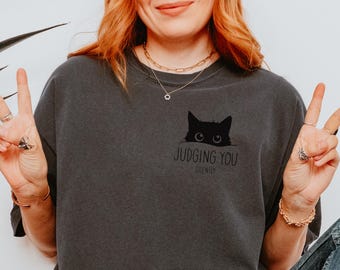 May include: A dark gray t-shirt with a black cat peeking over the top and the text "Judging You Silently".