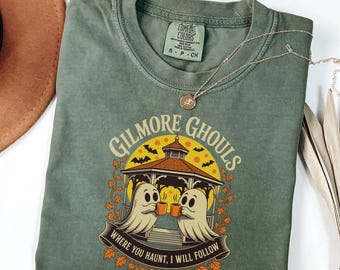 Pode incluir: Camiseta verde musgo com o texto "GILMORE GHOULS" e um gráfico de duas figuras de fantasmas segurando canecas sob um gazebo. A frase "WHERE YOU HAUNT, I WILL FOLLOW" está impressa abaixo. Um colar de prata está sobre a camiseta.