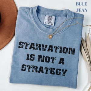 May include: A blue jean Comfort Colors t-shirt with the text "STARVATION IS NOT A STRATEGY" in black block letters. A gold necklace with a pendant is draped on the shirt.