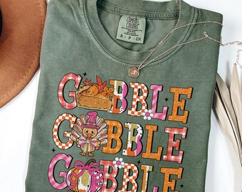 May include: A moss green Comfort Colors t-shirt with a Thanksgiving-themed graphic. The graphic says "Gobble Gobble" in a mix of orange, pink, and plaid patterns, with images of a pie, turkey, and pumpkin. A silver necklace is draped across the shirt.