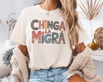 May include: Ivory-colored t-shirt with the text "CHINGA LA MIGRA" in colorful, decorative lettering. The shirt features a crew neck and short sleeves. The text is in a bold, eye-catching font with floral and geometric patterns.