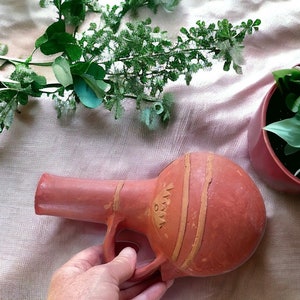 May include: A terracotta clay jug with a narrow spout and a handle. The jug has a decorative design of lines and a symbol on the side.
