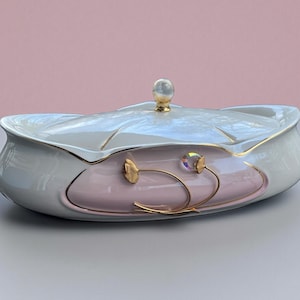 May include: Oval ceramic serving dish with a lid. The dish is primarily white with a pink center and gold accents. A clear, round knob tops the lid. Gold floral designs adorn the side, and a clear crystal-like accent is present.