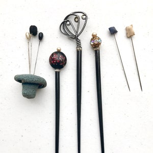 May include: A collection of decorative hat pins and hair pins. The pins feature various designs, including a crochet hat, pearl and bead accents, and jeweled toppers. The pins are in various colors, including black, silver, and gold.