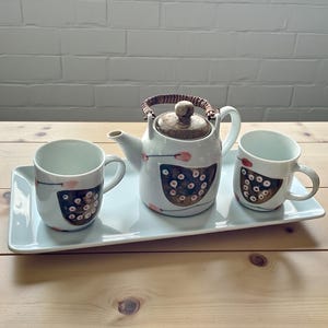 May include: A white ceramic tea set with a teapot, two mugs, and a rectangular tray. The teapot has a wooden lid and handle. The mugs and teapot are decorated with a brown and pink pattern. The set is on a wooden table.