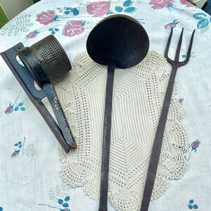 May include: Three vintage kitchen utensils: a black metal potato ricer, a black metal ladle, and a black metal fork. The utensils are arranged on a white crocheted doily on a white tablecloth with a floral pattern.